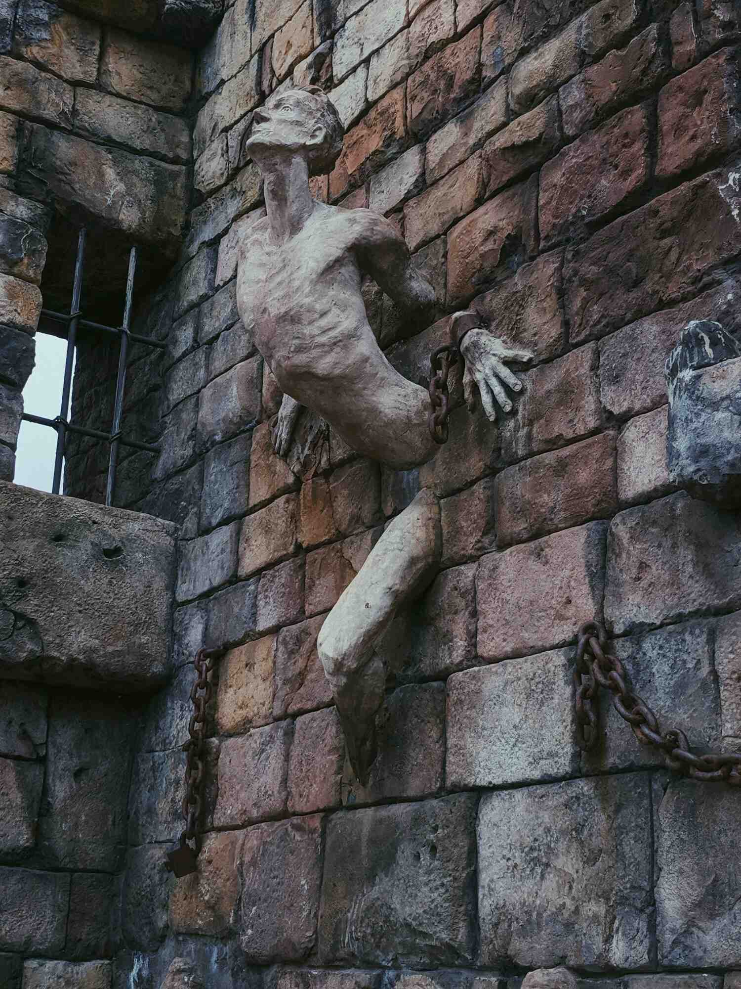 A side-view of a sculpture of an elderly man emerging from a wall, with shackles on his left wrist and chains along the bricks as he looks outwards, with a barred window in the corner brick wall gap in the foreground