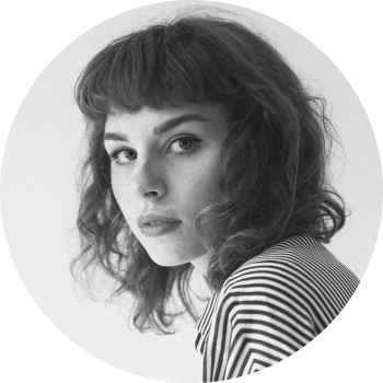 A black and white headshot of a young girl in her mid-twenties with dark, curly hair and a striped top staring at the camera positioned sideways